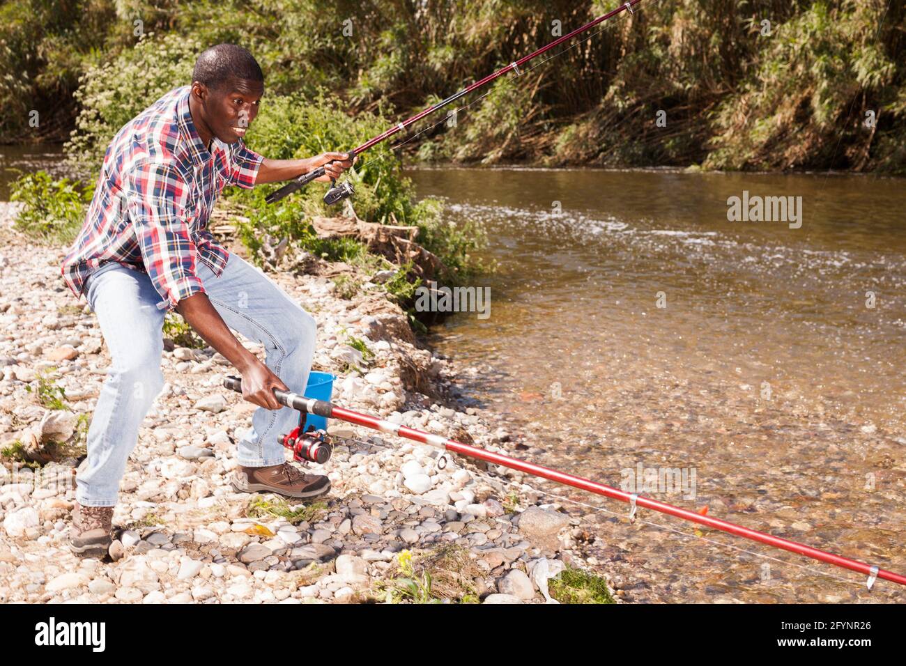 Portrait of enthusiastic African man pulling fish with two rods on river Stock Photo