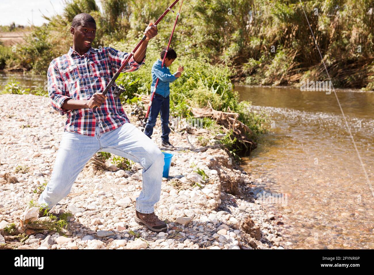 Portrait of Afro man pulling fish with fishing rod and his son fishing on river Stock Photo