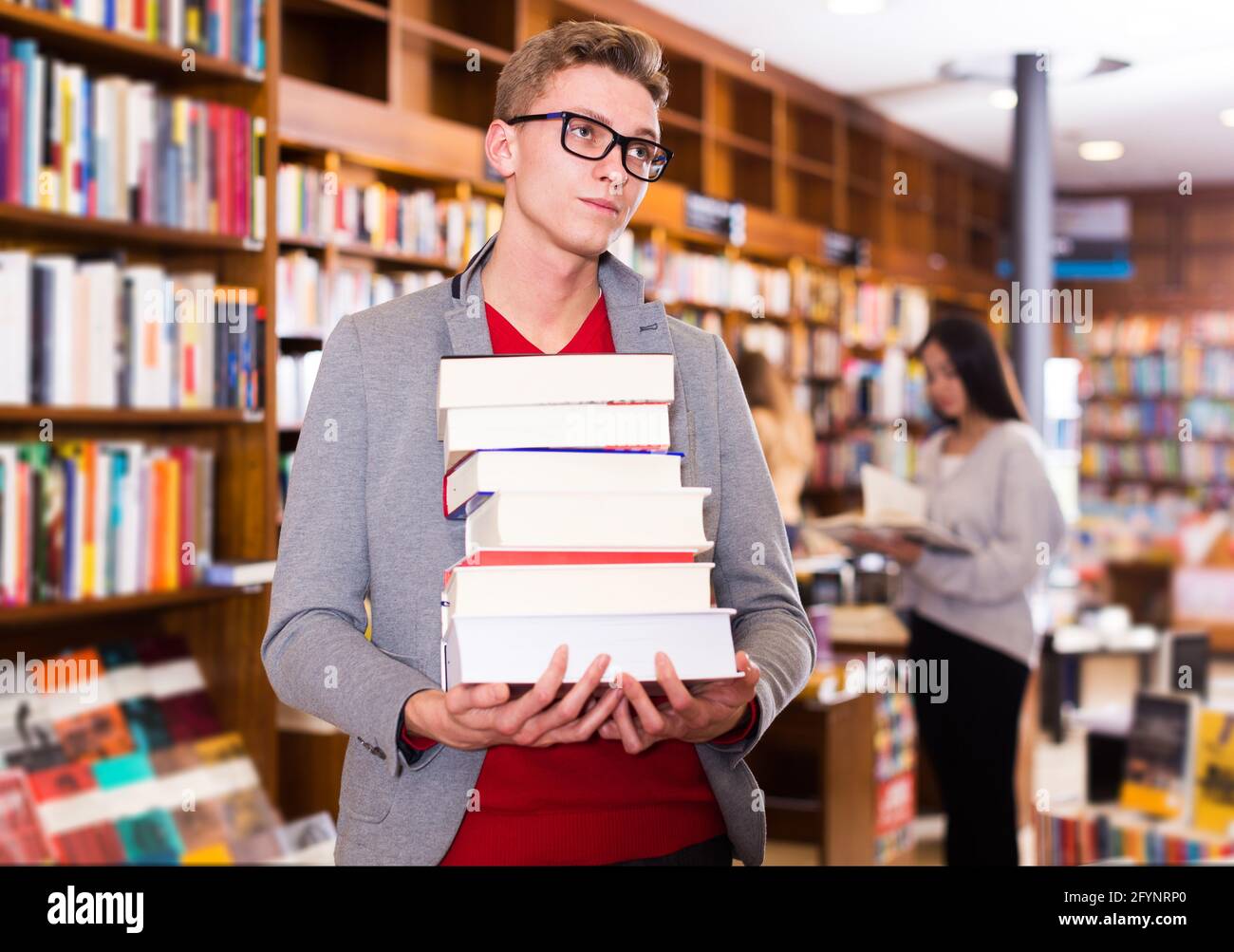 Tired young student standing in library with pile of books in hands ...