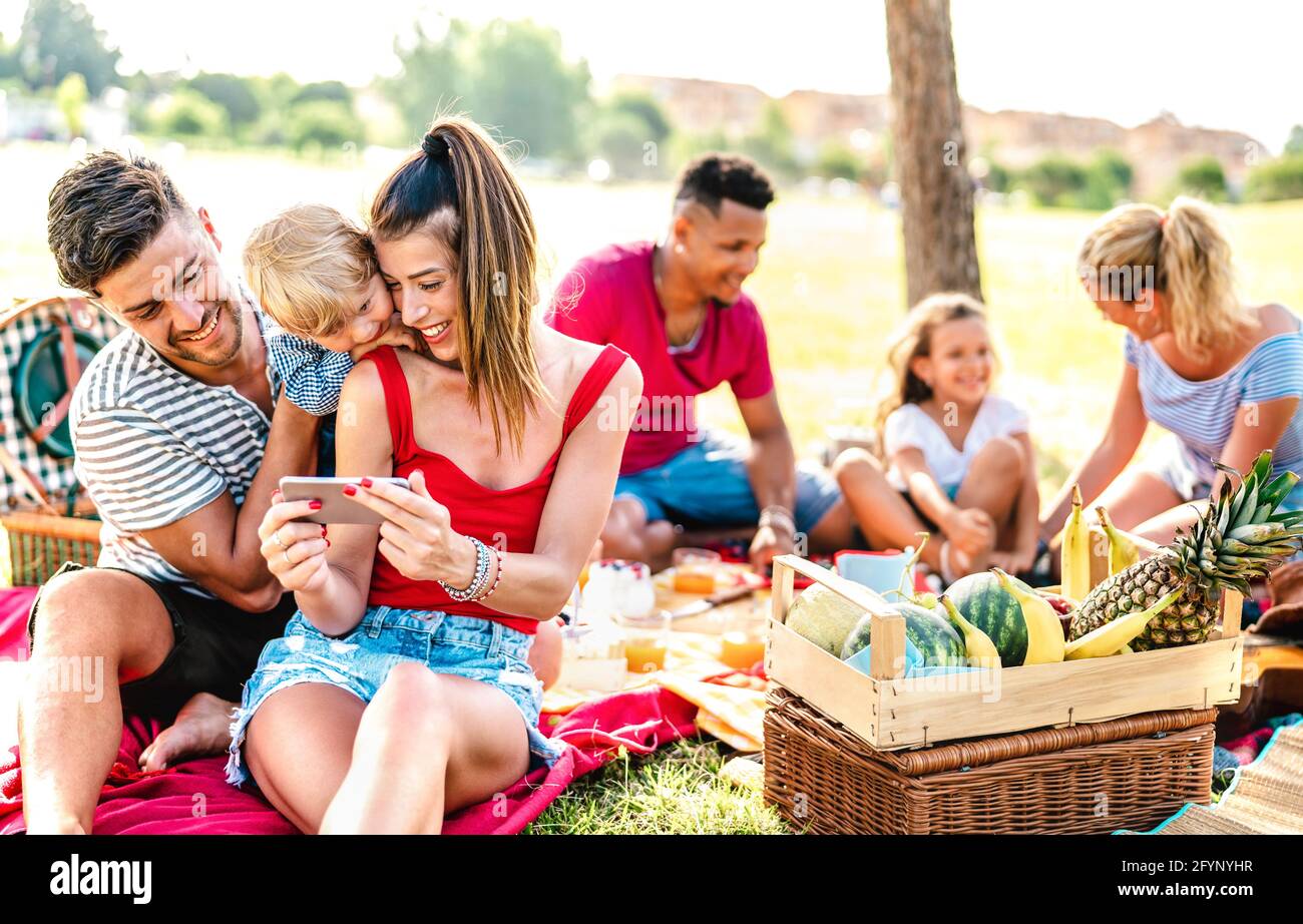 Happy multiethnic families playing with phone at pic nic garden party - Joy and love life style concept with mixed race people having fun together Stock Photo