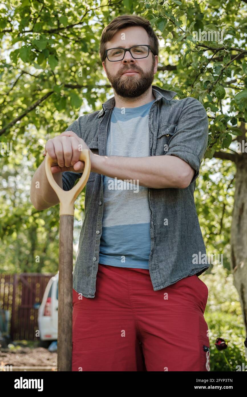 Serious man is leaning on the handle of a shovel and looking at the camera, in the garden, against the backdrop of a car and a fence. Stock Photo