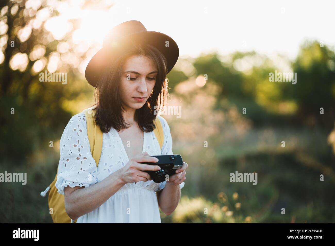 Fashion look, pretty cool young woman model with retro film camera and wearing a elegant hat, white dress, yellow backpack. Female tourist holding vin Stock Photo