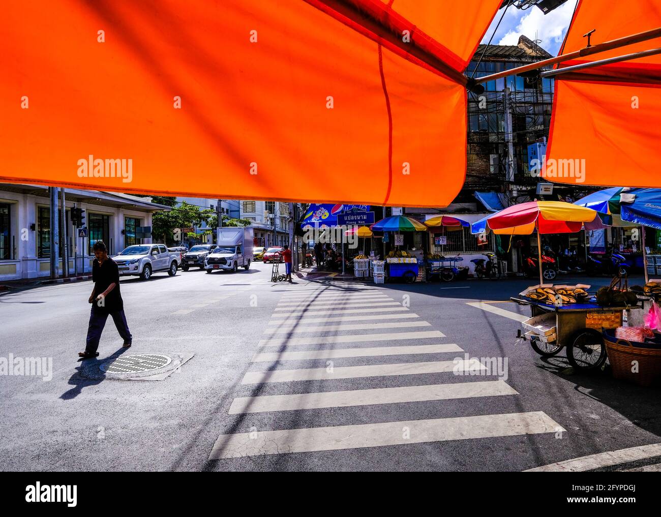 A man crosses a sunlit road against a backdrop of colorful parasols shielding various street vendors from the sun in Chinatown, Bangkok, Thailand. Stock Photo