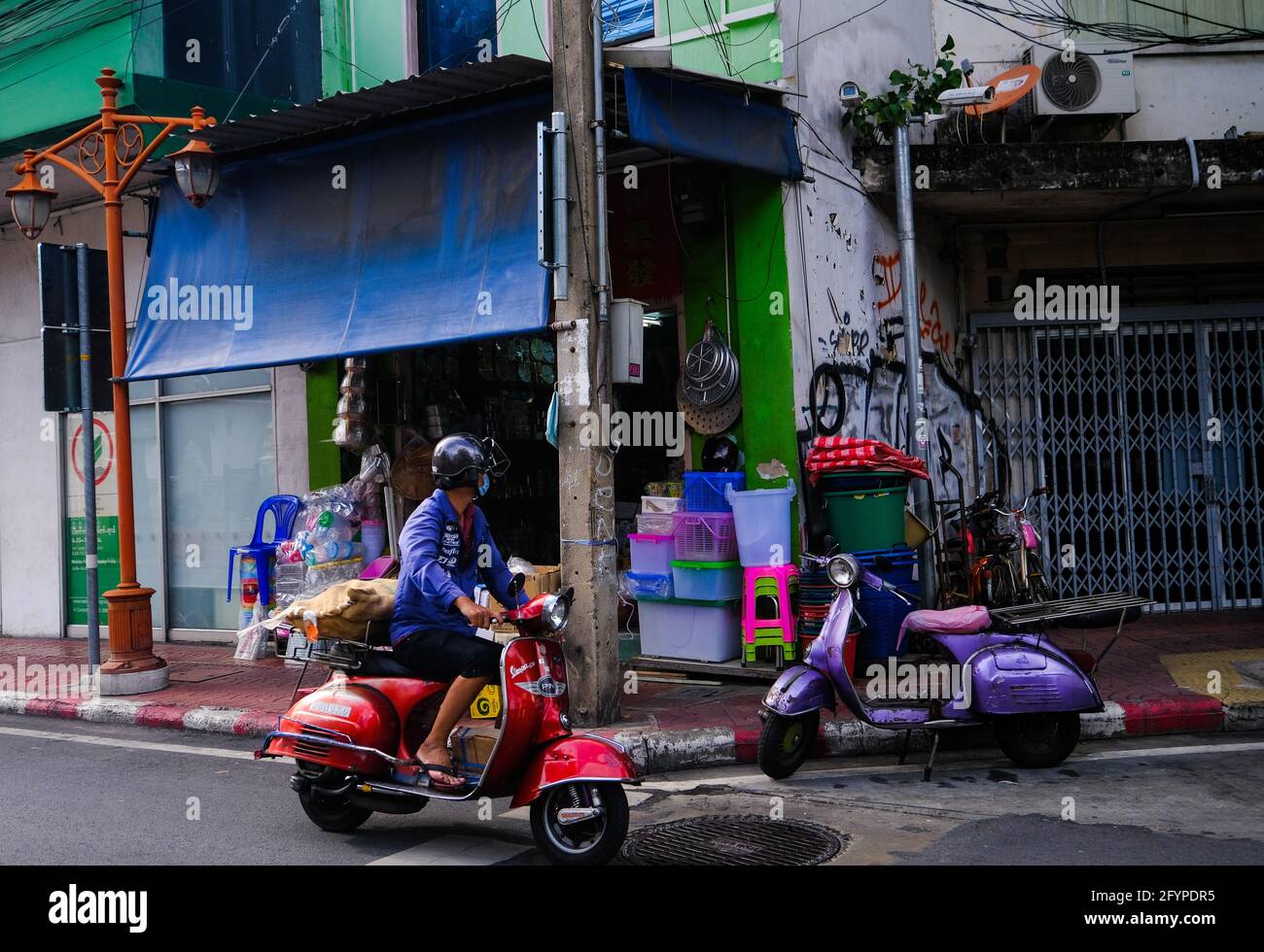A man riding a red Vespa scooter goes past a colorful shopfront in Talat Noi, Bangkok, Thailand Stock Photo