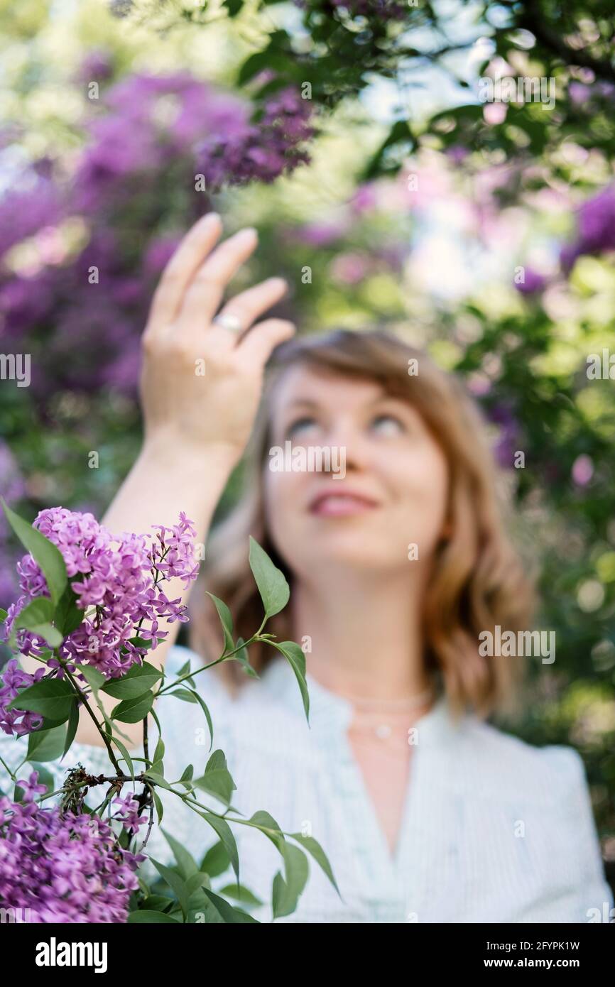 Candid authentic portrait of 30s 40s caucasian blonde woman with lilac flowers. 30 40 year old woman enjoying life in lilac flowers nature background Stock Photo