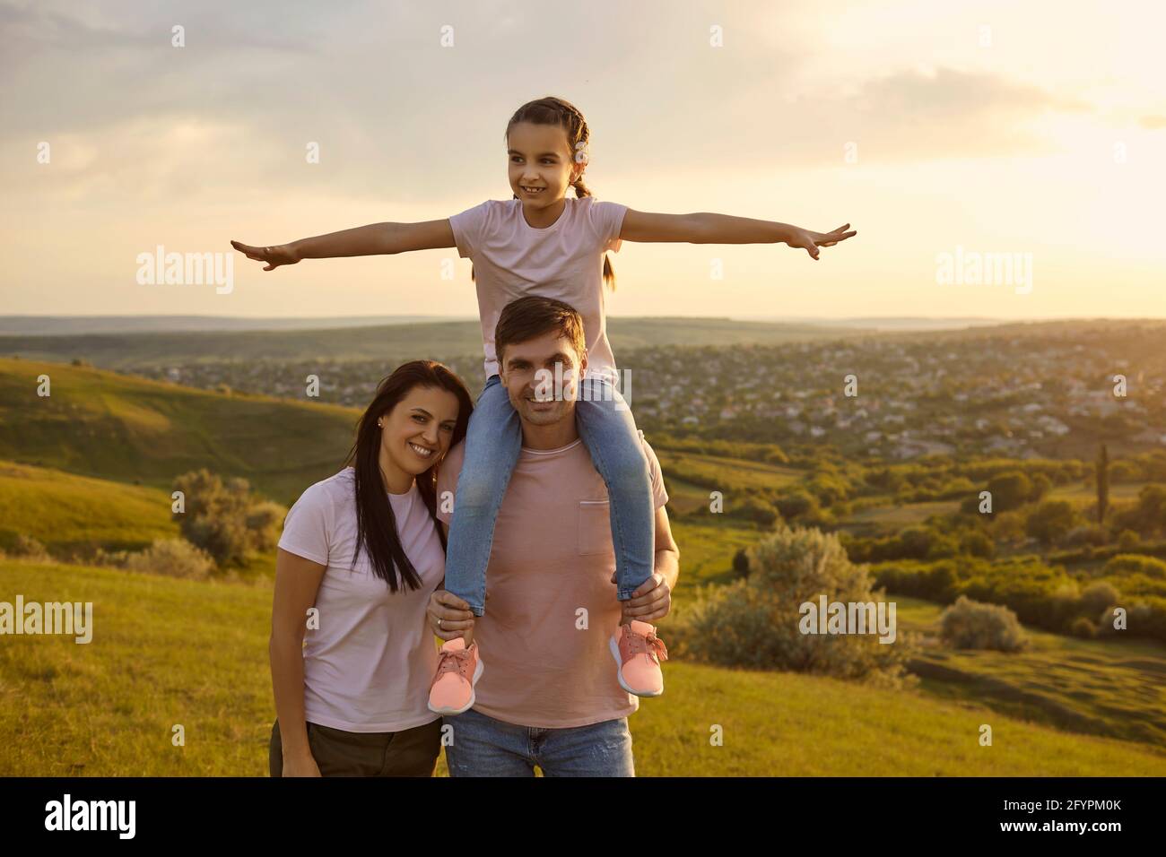 Daughter on the shoulders of parents smiling walk on the grass at sunset in nature. Stock Photo