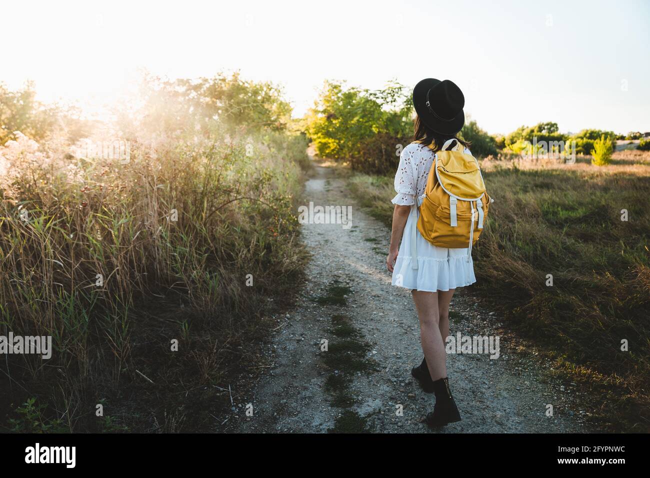 Back view of female backpacker walking on forest path and viewing landscape in summer vacation. Stylish young woman with hat and white dress exploring Stock Photo