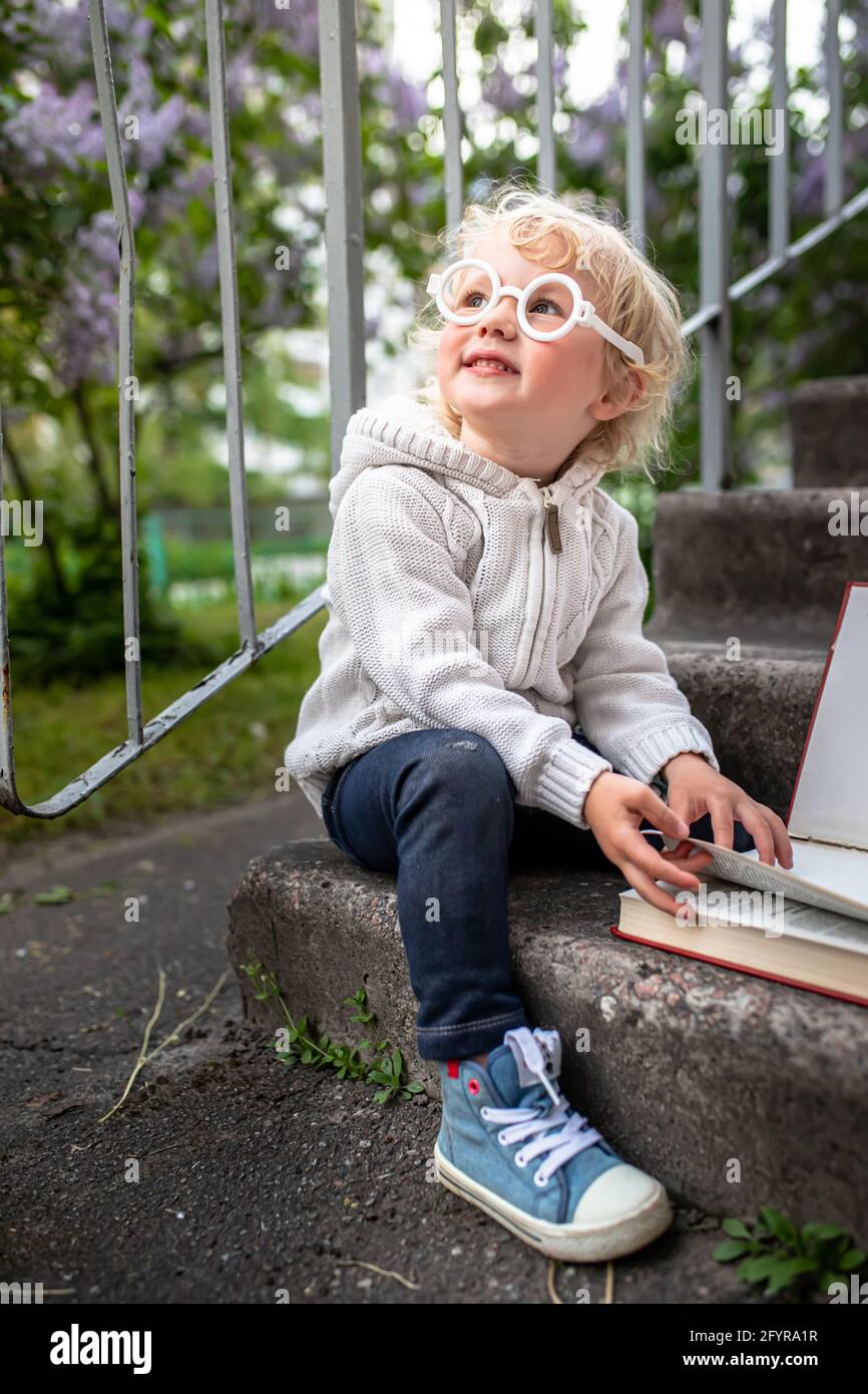 A girl with glasses sits next to an open book. Pre-school education. Funny baby in a knitted jacket and sneakers Stock Photo