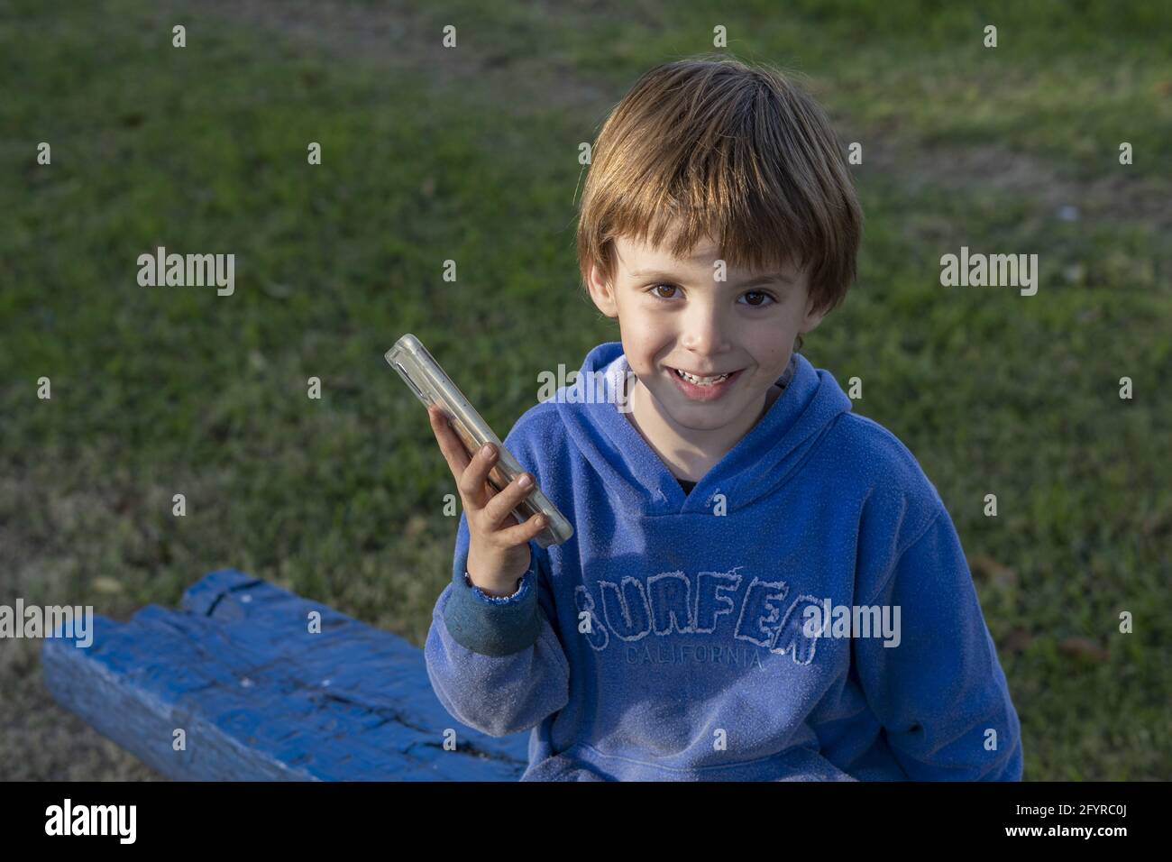 Caucasian boy with a blue hoodie sitting on bench in park and holding a  phone with a smiley face Stock Photo - Alamy