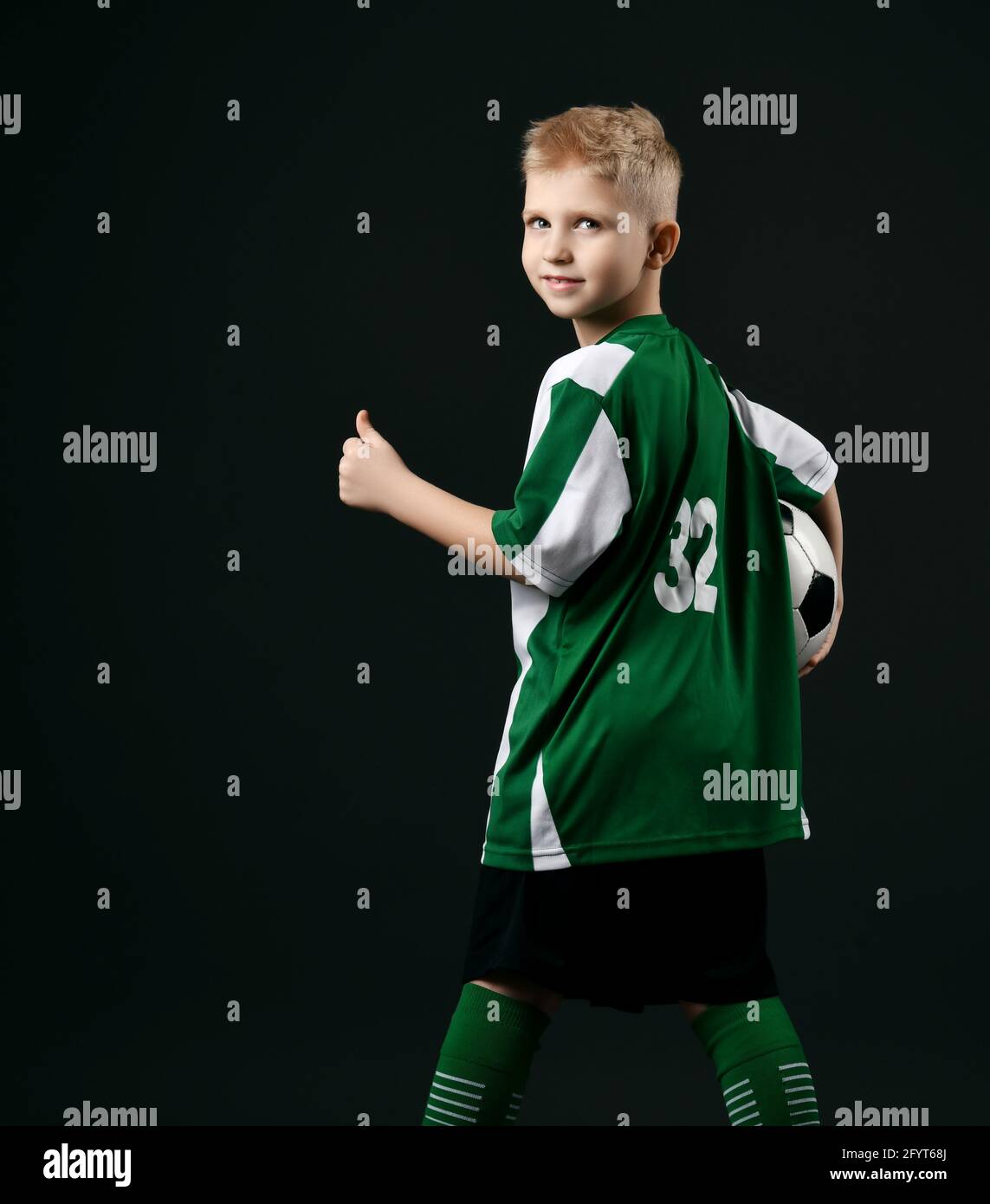 Blond smiling boy child in sports green and white clothes standing holding soccer ball and showing thumbs up sign Stock Photo