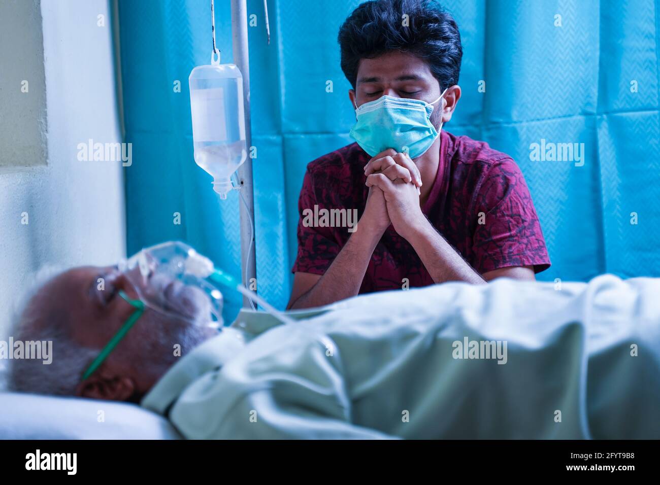 Worried stressed out young man praying for his father to recover from covid 19 infection while breathing on ventilator oxygen mask at hospital - Stock Photo