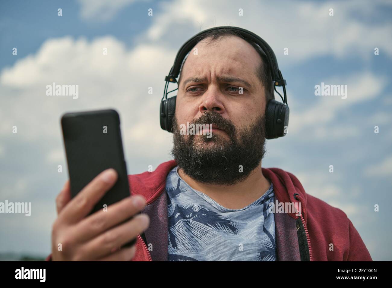 Middle-aged European man in headphones outdoors listening to music against the background of the sky, mobile phone in his hand Stock Photo
