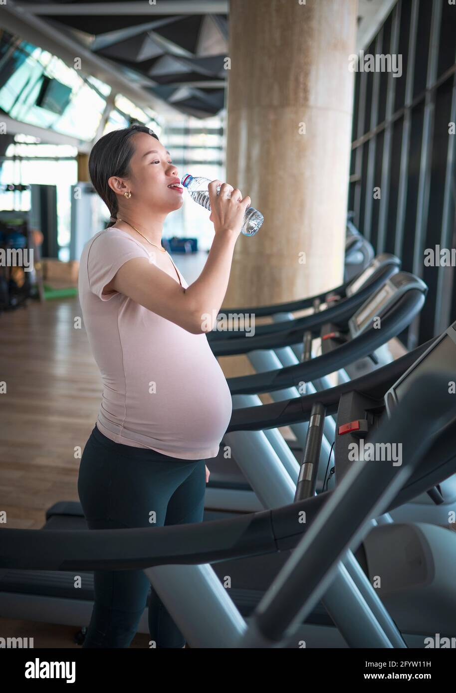 Pregnant Asian woman hydrating while exercising in the gym indoors walking on a treadmill for a fit and active lifestyle during pregnancy Stock Photo