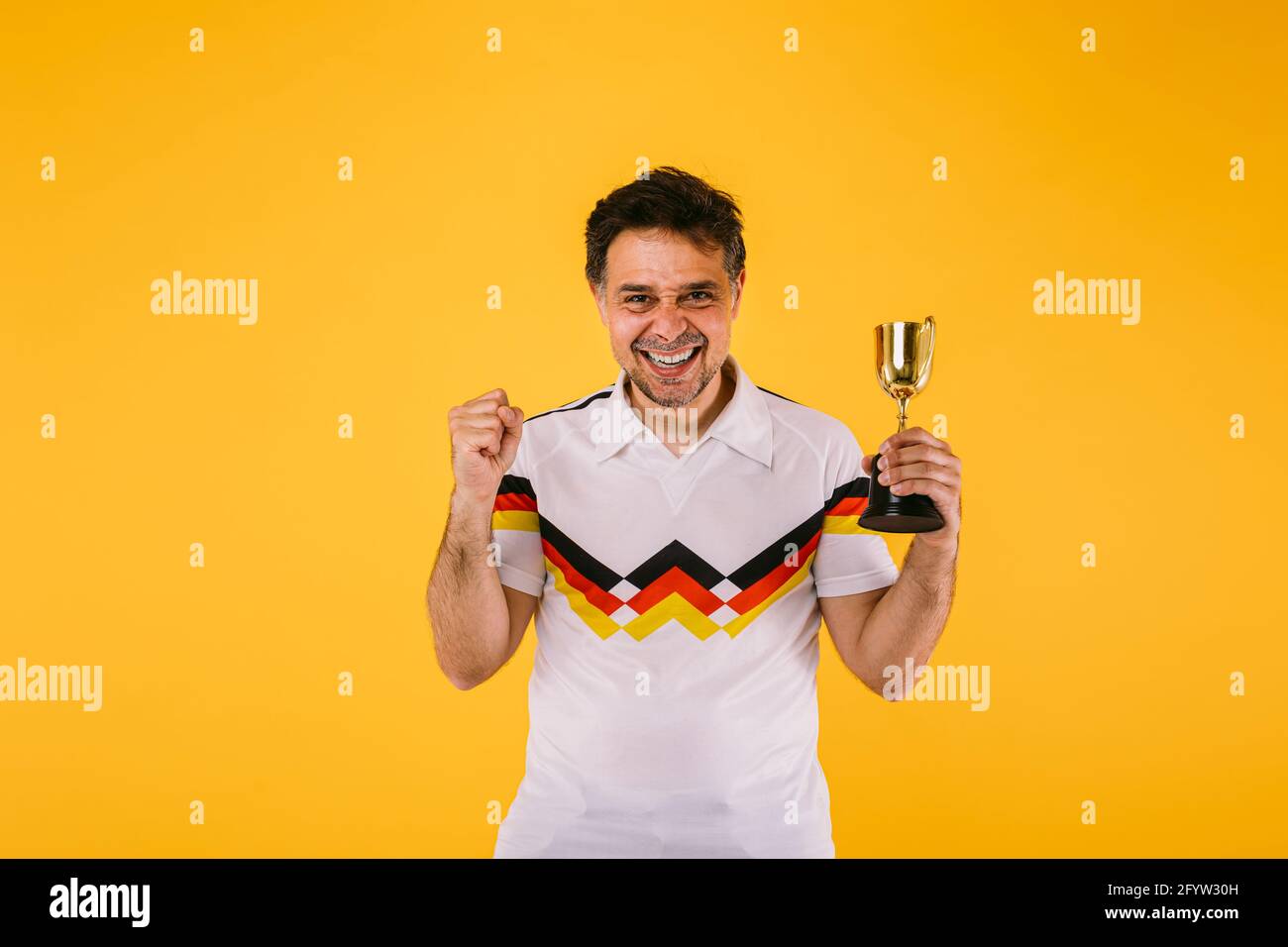 Soccer fan wearing a white t-shirt with black, red and yellow stripes, he clenches his fist and holds a winner's trophy. Stock Photo