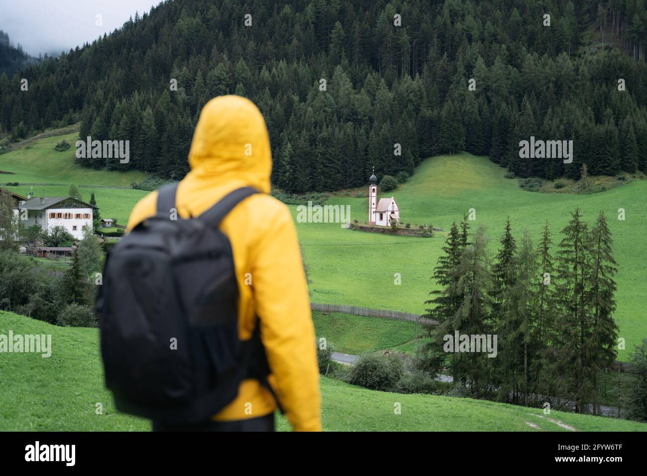 Hiking man with backpack enjoy the view of St. Johann church in Val di Funes valley, Dolomites, Italy, Europe Stock Photo