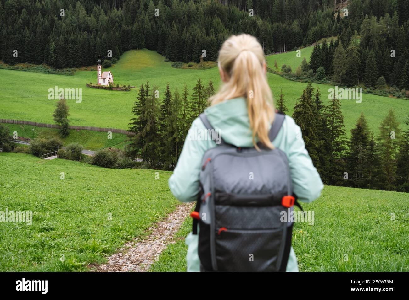Hiking woman with backpack enjoy the view of St. Johann church in Val di Funes valley, Dolomites, Italy, Europe Stock Photo