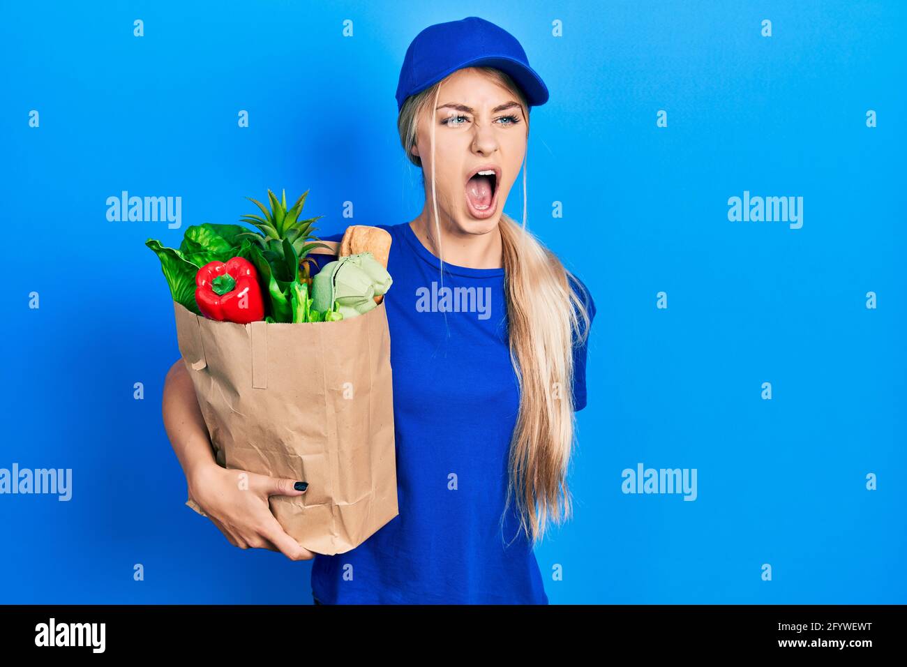 Young caucasian woman wearing courier uniform with groceries from supermarket angry and mad screaming frustrated and furious, shouting with anger. rag Stock Photo