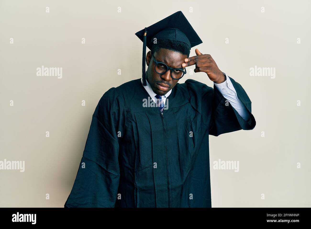 Handsome black man wearing graduation cap and ceremony robe pointing unhappy to pimple on forehead, ugly infection of blackhead. acne and skin problem Stock Photo