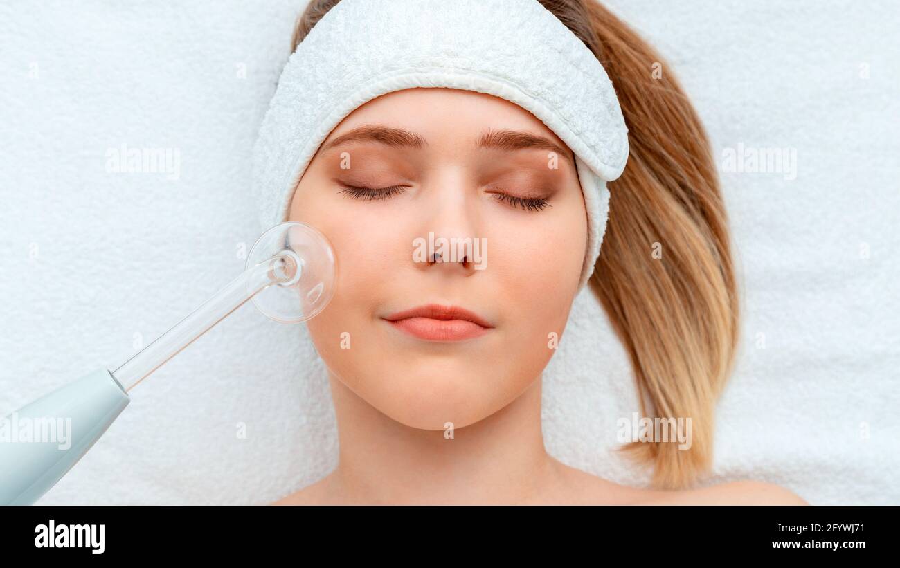 Woman client portrait during darsonval cosmetology procedure for skin care health lies on white towel. Cosmetics and beautician tool device for face Stock Photo