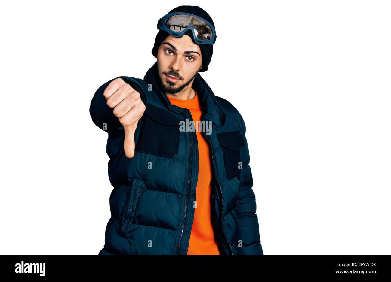 Young hispanic man with beard wearing snow wear and sky glasses looking unhappy and angry showing rejection and negative with thumbs down gesture. bad Stock Photo
