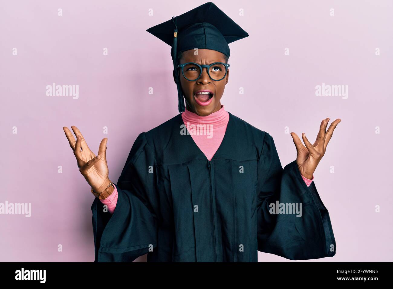 Young african american girl wearing graduation cap and ceremony robe crazy and mad shouting and yelling with aggressive expression and arms raised. fr Stock Photo