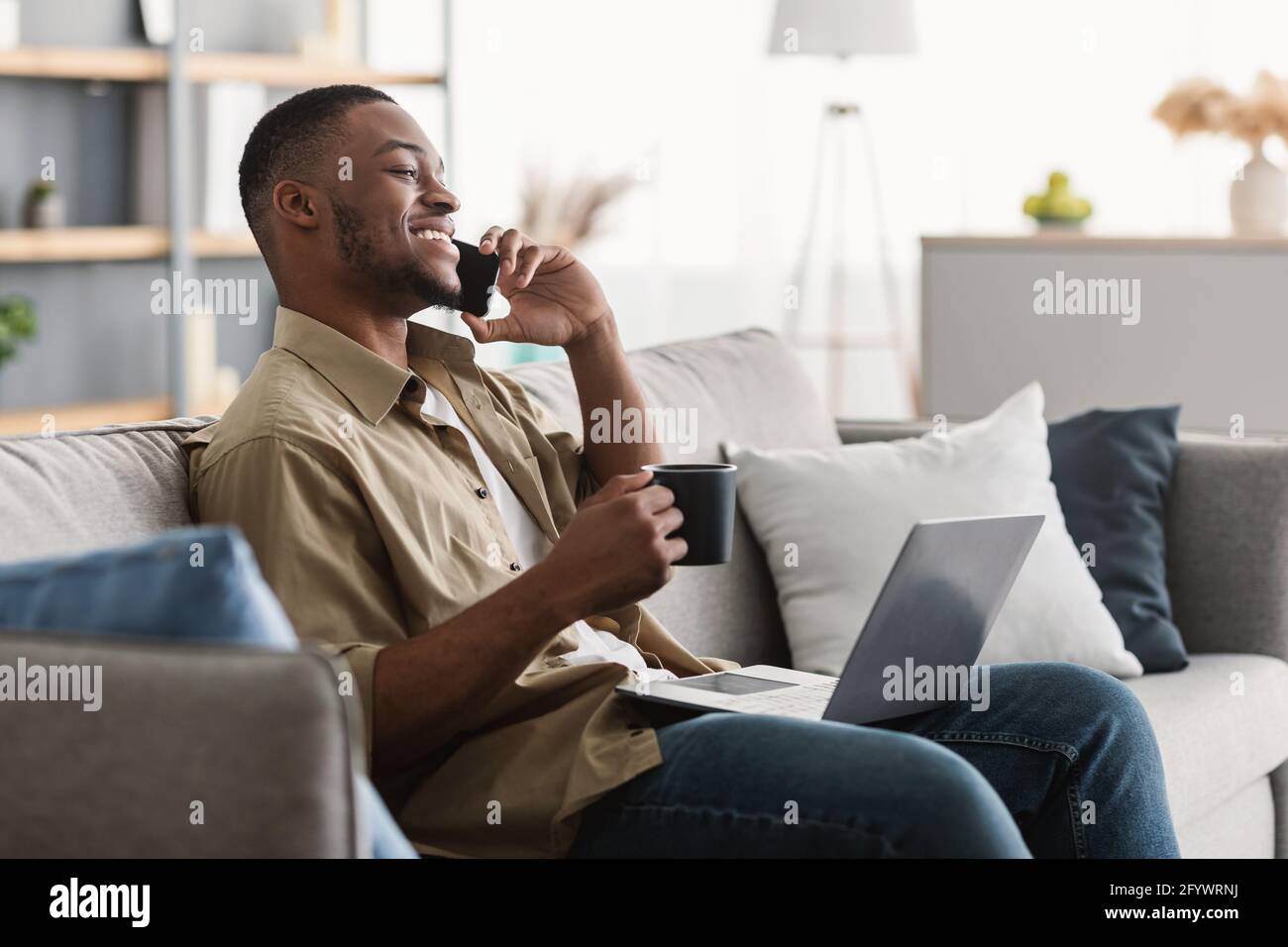 Black Man Talking On Cellphone Using Laptop Drinking Coffee Indoors Stock Photo