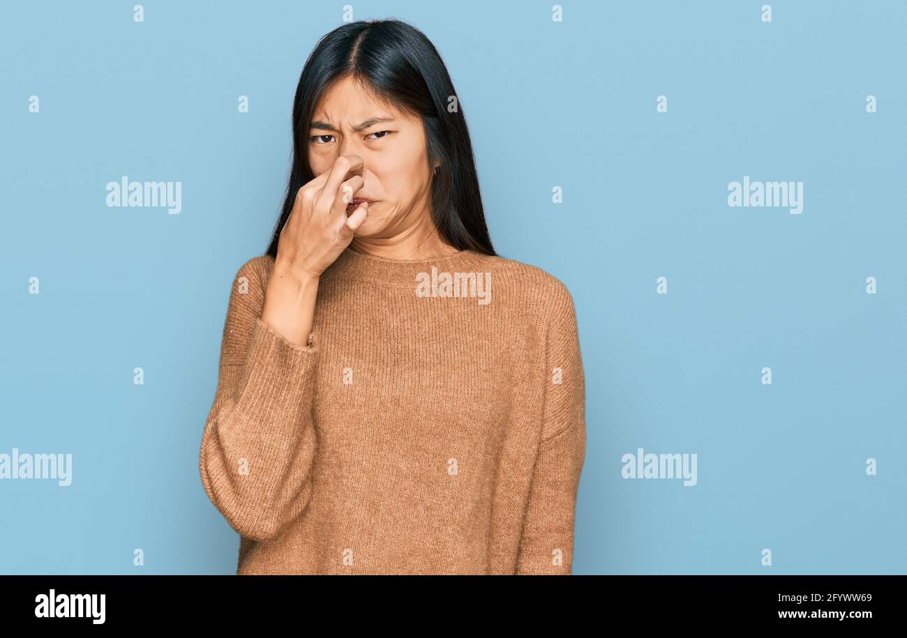 Beautiful young asian woman wearing casual winter sweater smelling something stinky and disgusting, intolerable smell, holding breath with fingers on Stock Photo