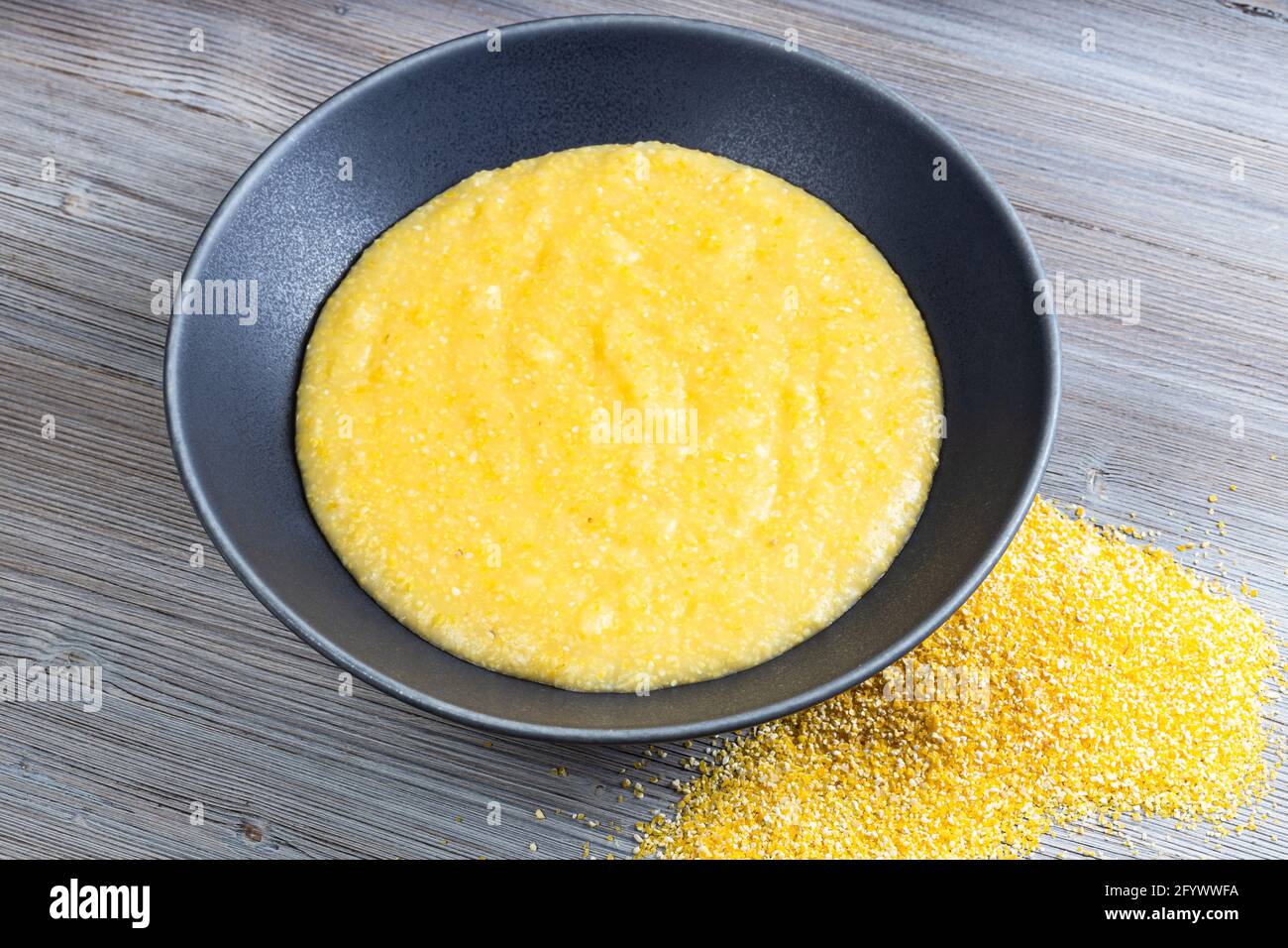 cooked maize porridge in gray bowl and handful of cornmeal on gray wooden table Stock Photo