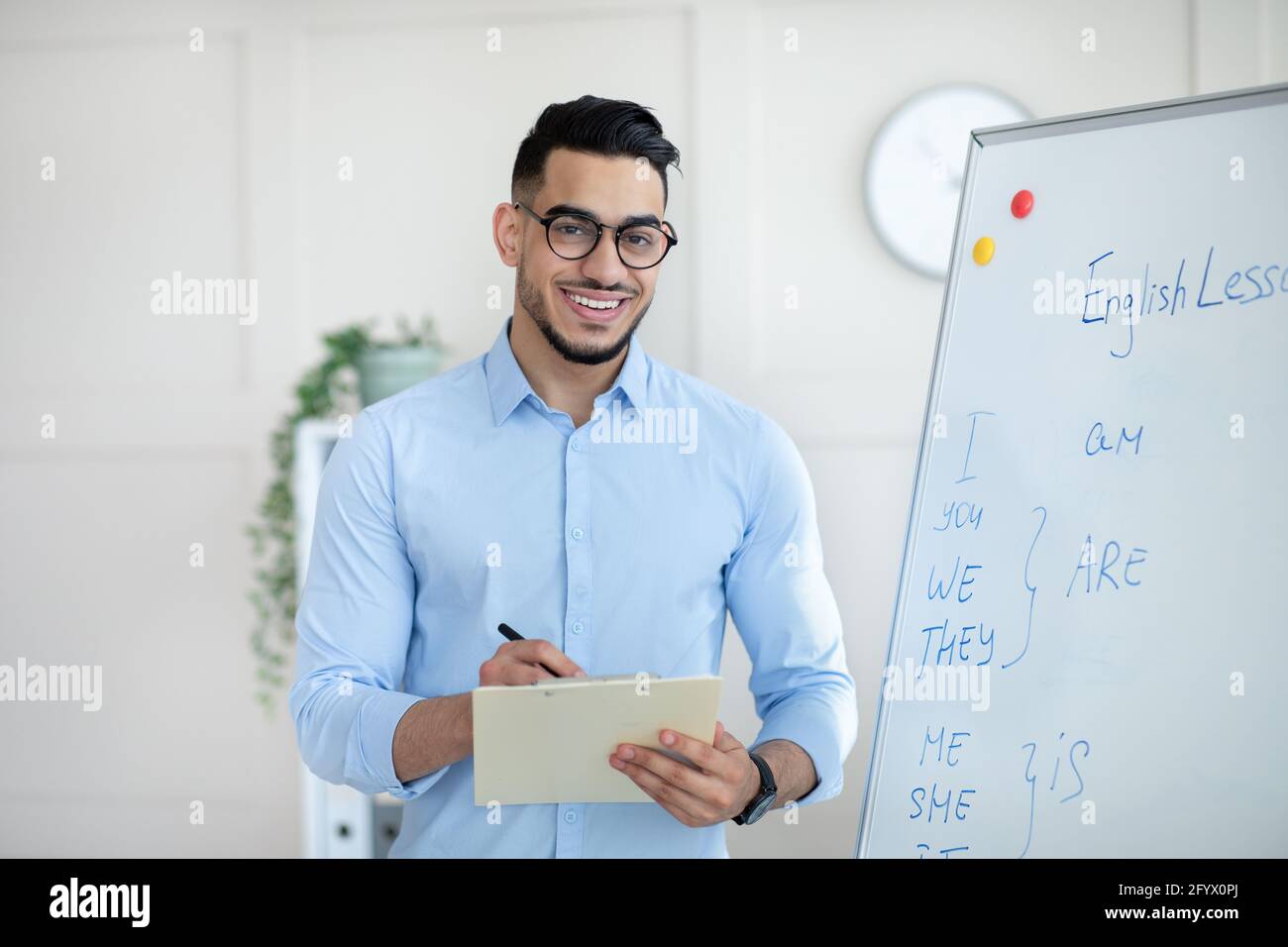 Positive male English teacher explaining foreign language rules near blackboard, taking notes in clipboard at office Stock Photo