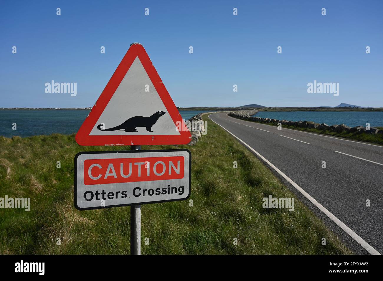 Caution Otters Crossing sign, red warning triangle and otter icon. Blurred background of causeway, sea and grass. South Uist to Benbecula causeway. Stock Photo