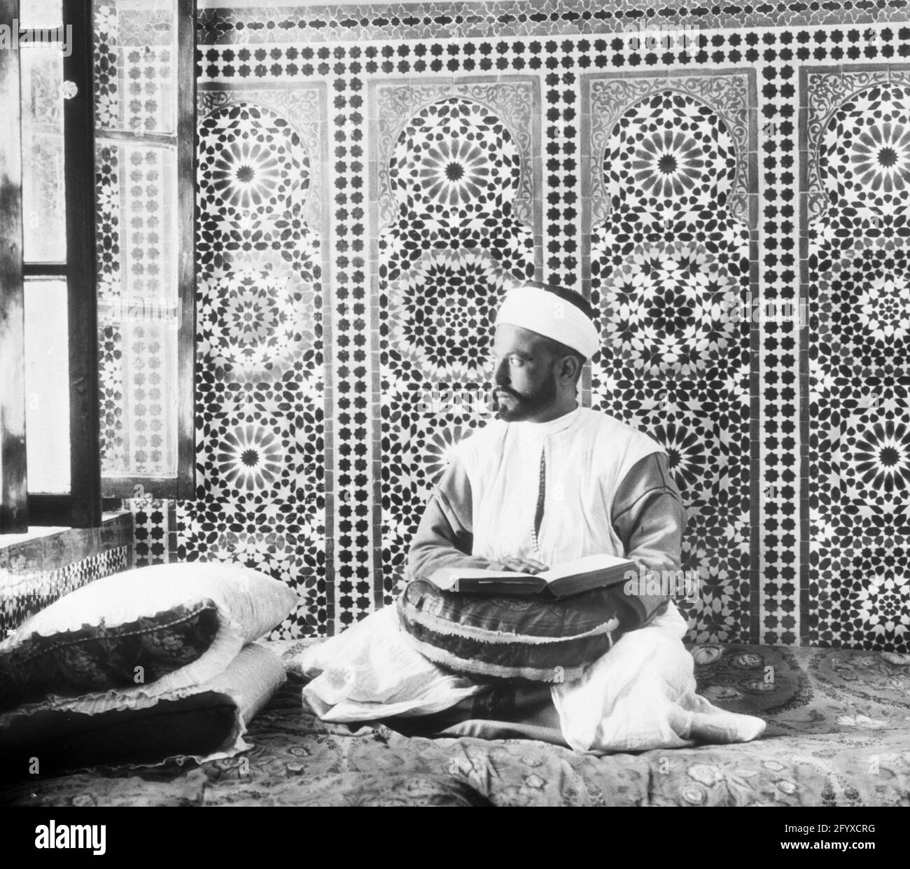 A man seated on a carpet near an open window reads the Quran, Fez, Morocco, 1923. (Photo by Burton Holmes) Stock Photo