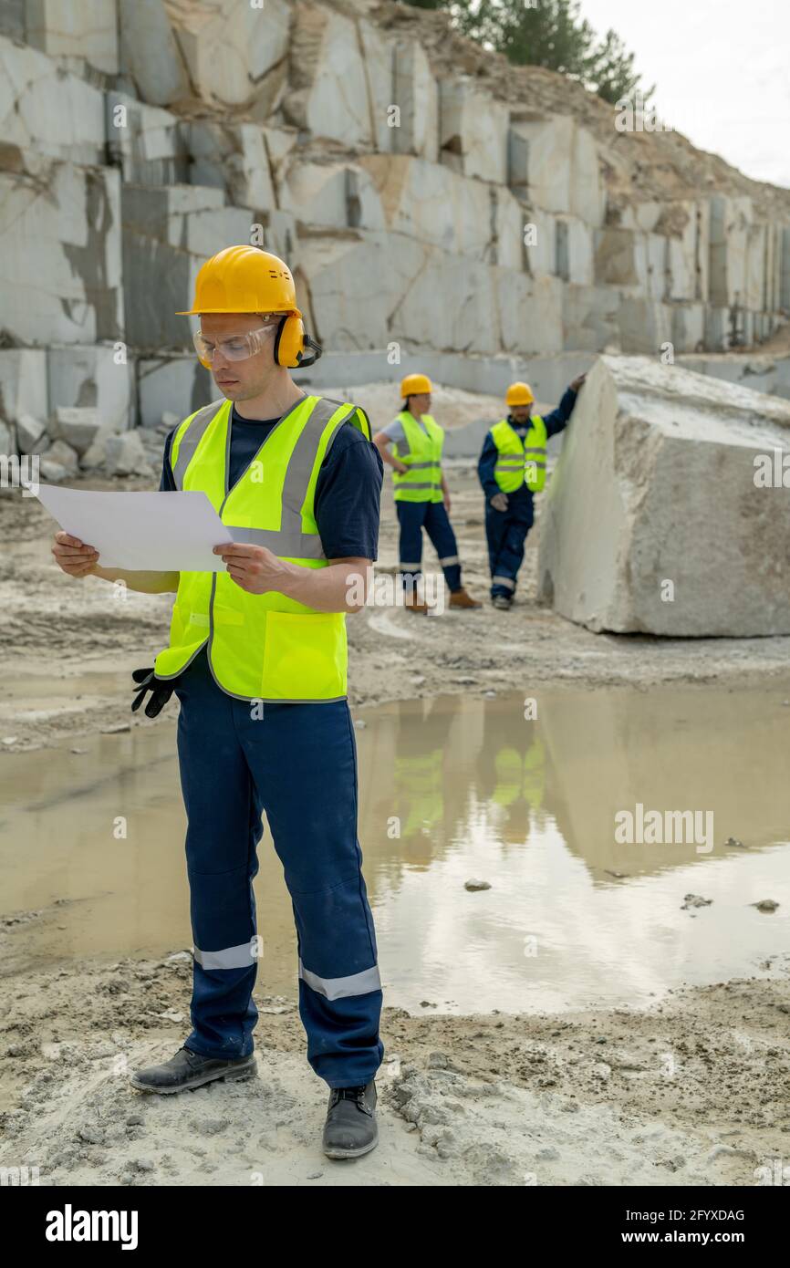 Young builder with sketch standing on construction site against two colleagues Stock Photo