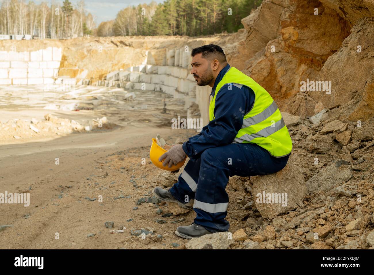 Middle aged builder in uniform sitting on stone while having break Stock Photo