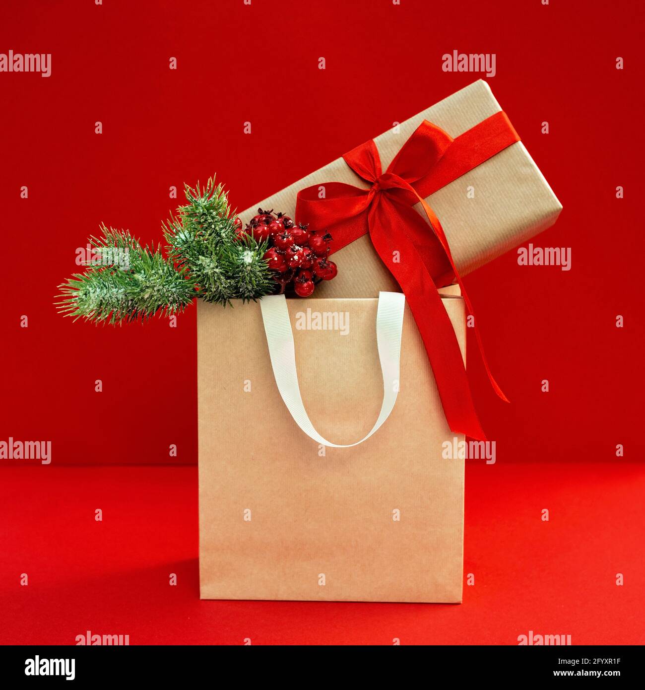 Gift bag with decorated kraft box with a branch of a Christmas tree on a red background. Preparation, shopping of gifts, sale in the New Year and Chri Stock Photo