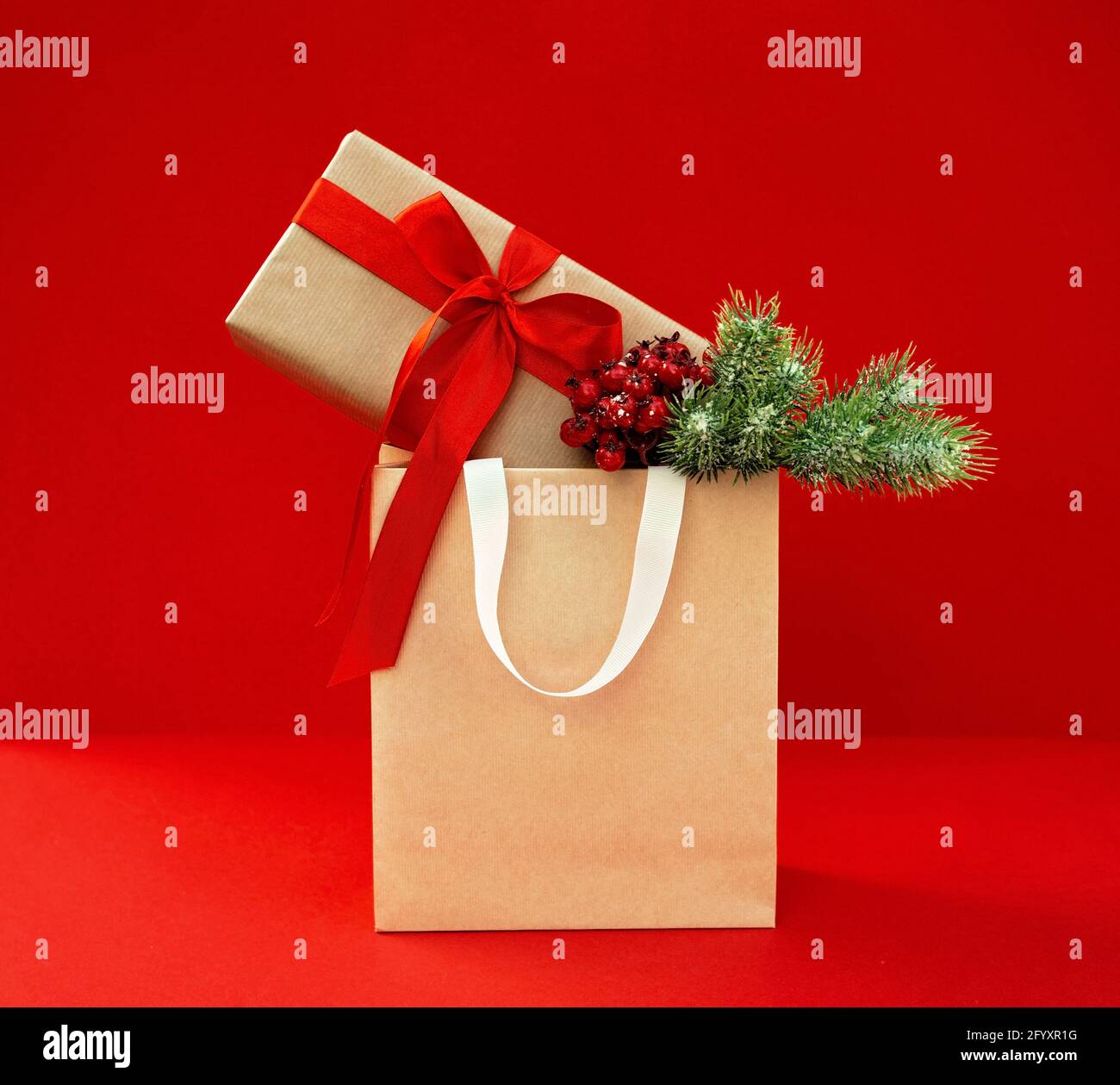 Gift bag with decorated kraft box with a branch of a Christmas tree on a red background. Preparation, shopping of gifts, sale in the New Year and Chri Stock Photo