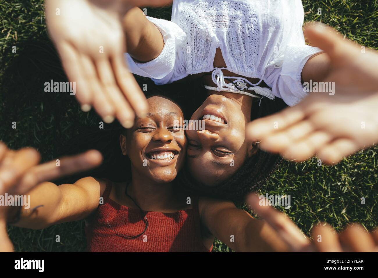 Top view of smiling African American female friends reaching hands to camera while lying on grassy lawn on sunny summer day Stock Photo