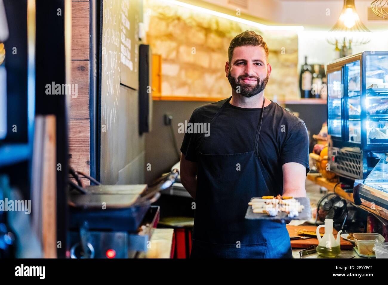 Happy ethnic male with beard demonstrating tray with tasty food while looking at camera in restaurant Stock Photo