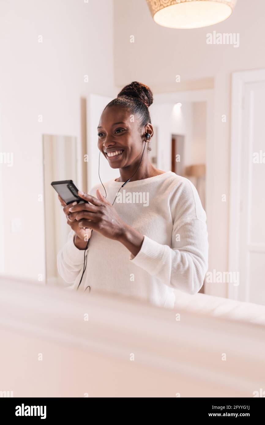 Happy young African American lady in casual clothes smiling while enjoying music in earphones with smartphone in hands Stock Photo
