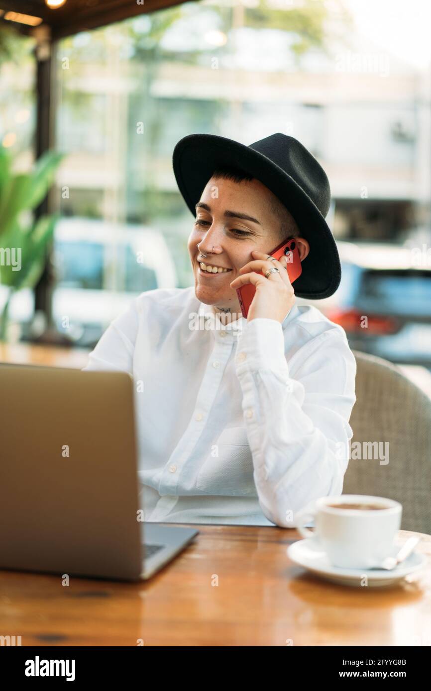 Stylish happy female freelancer having phone call while working in cafe with laptop with cup of coffee Stock Photo