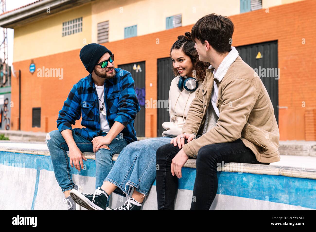 Cheerful friends speaking with each other and smiling on concrete parapet near brick building Stock Photo