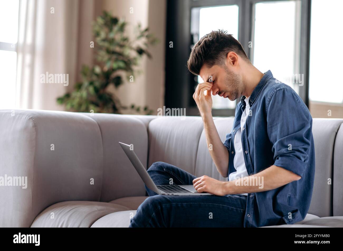 Upset tired young caucasian bearded man sitting on sofa in living room, in casual clothes, using laptop, student in stress, overworked with study and homework, rubs eyes, needs rest Stock Photo