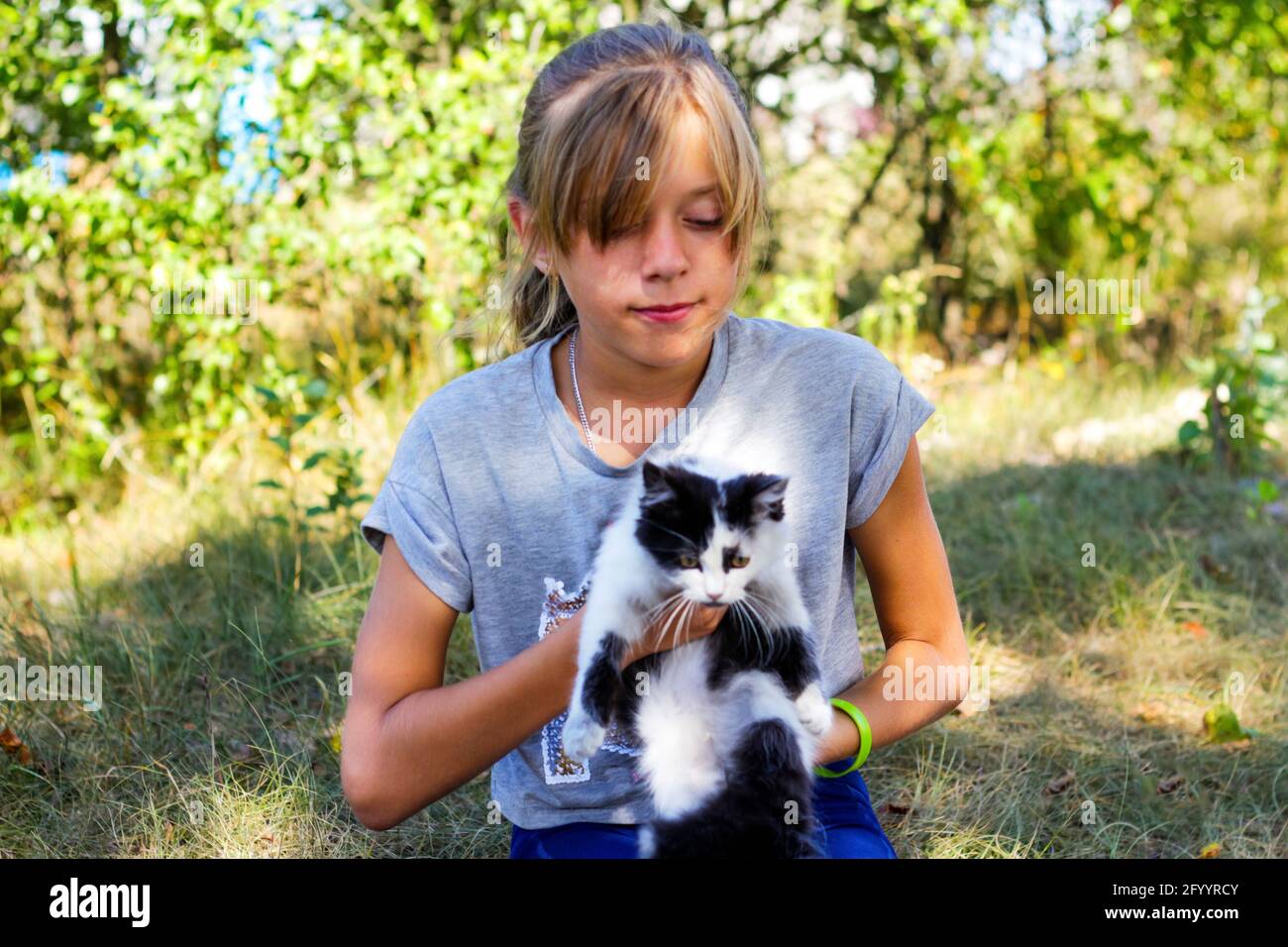 Defocus blonde little smiling girl playing with cat, black and white small kitten. Nature blurred green summer background. Girl holding and stroking p Stock Photo