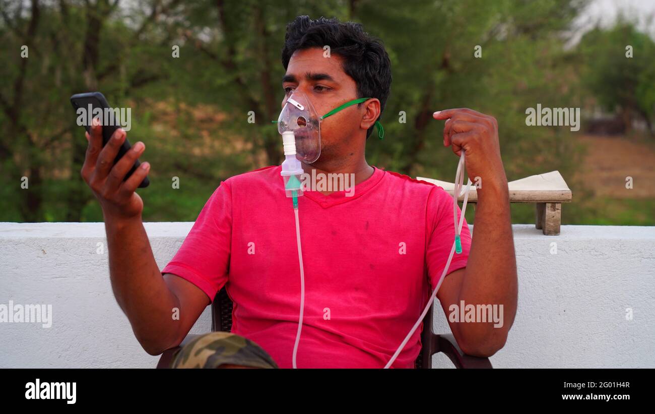 Young man infected with Covid 19 disease. Patient inhaling oxygen wearing mask and calling on mobile phone. liquid Oxygen flow in Rajasthan, India. Stock Photo