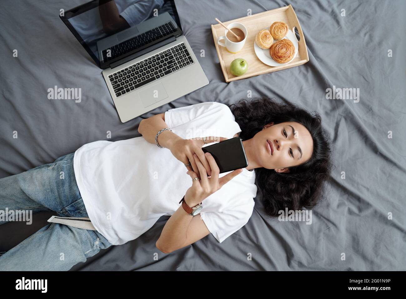 Directly above view of relaxed young Vietnamese guy with black hair lying on bed with laptop and tray with breakfast and exchanging messages on phone Stock Photo