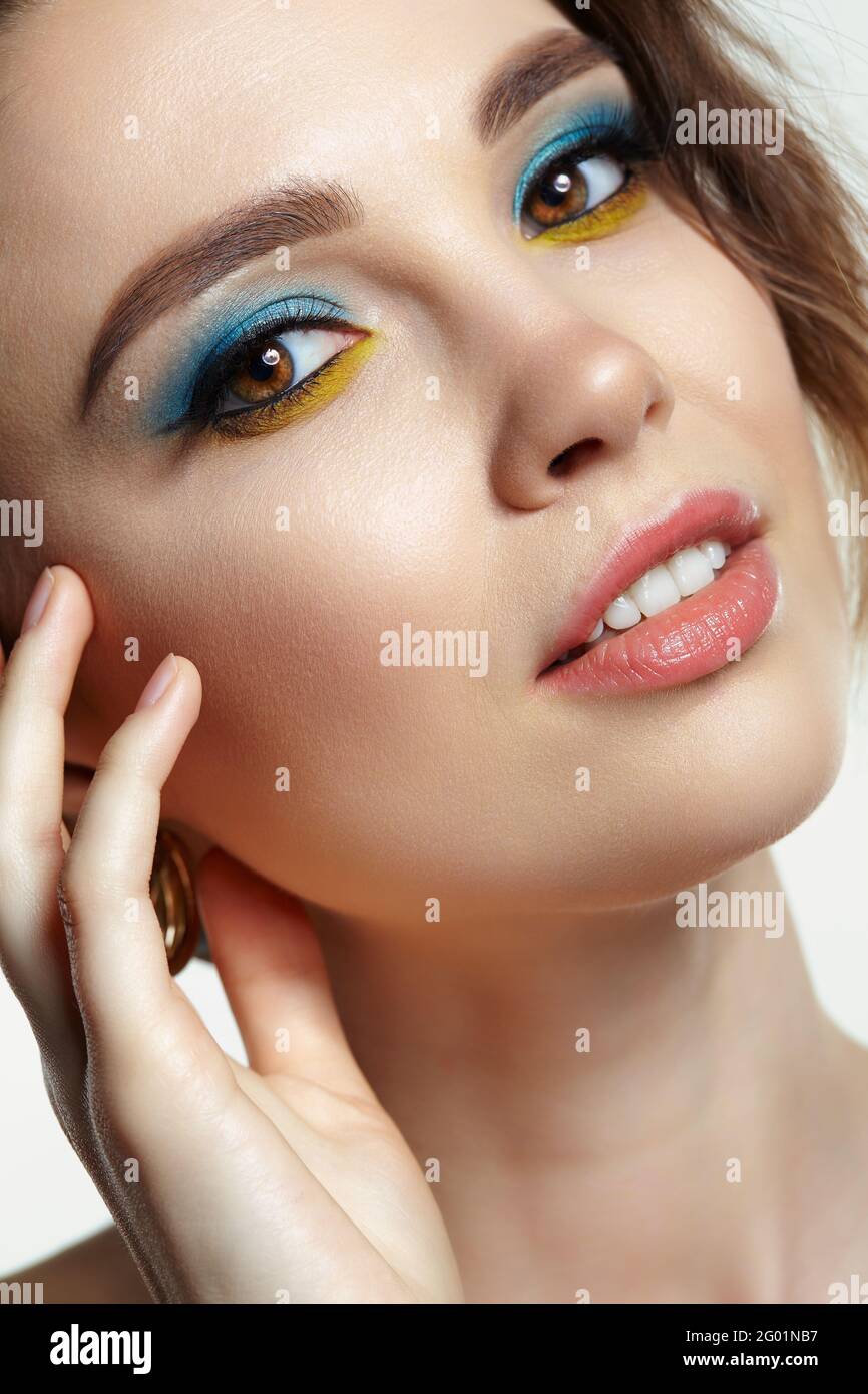 Portrait of young woman on gray background. Female posing with hand near face. Makeup with blue eyeshadow and yellow eyeliner. Stock Photo