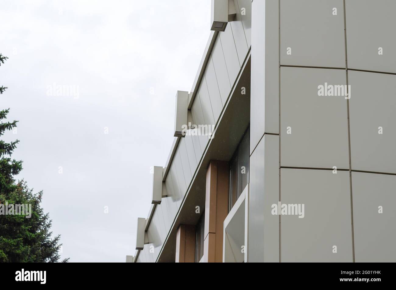 The beige and brown facade of a modern building against an overcast sky. On the left is a tall green fir tree. Daytime. Shooting in the rain. Architec Stock Photo