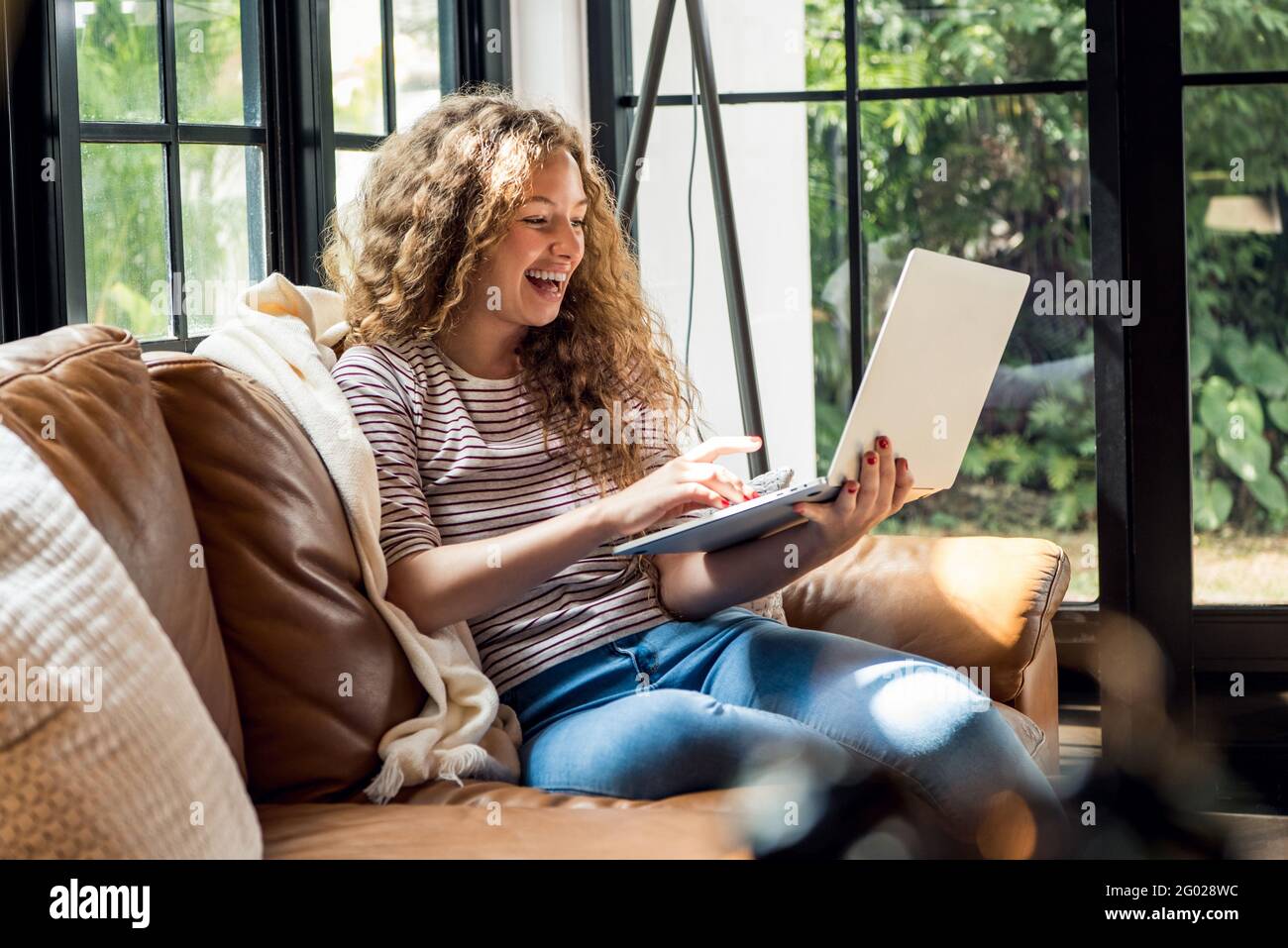 Happy smiling caucasian woman chatting via video call with laptop computer on the couch in living room at home during day time Stock Photo