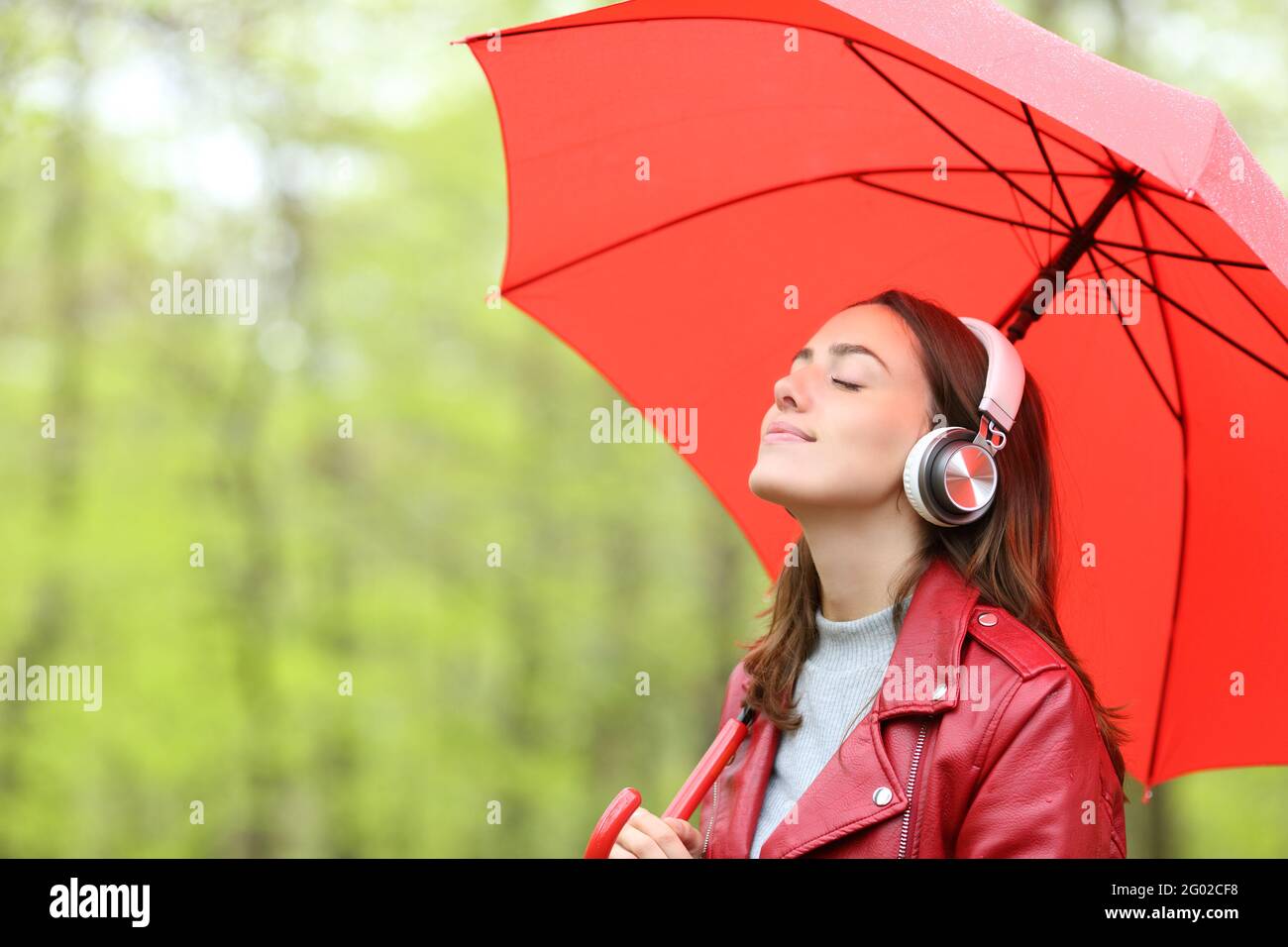 Relaxed woman in red holding umbrella under the rain listening to music wearing wireless headphones in a park Stock Photo