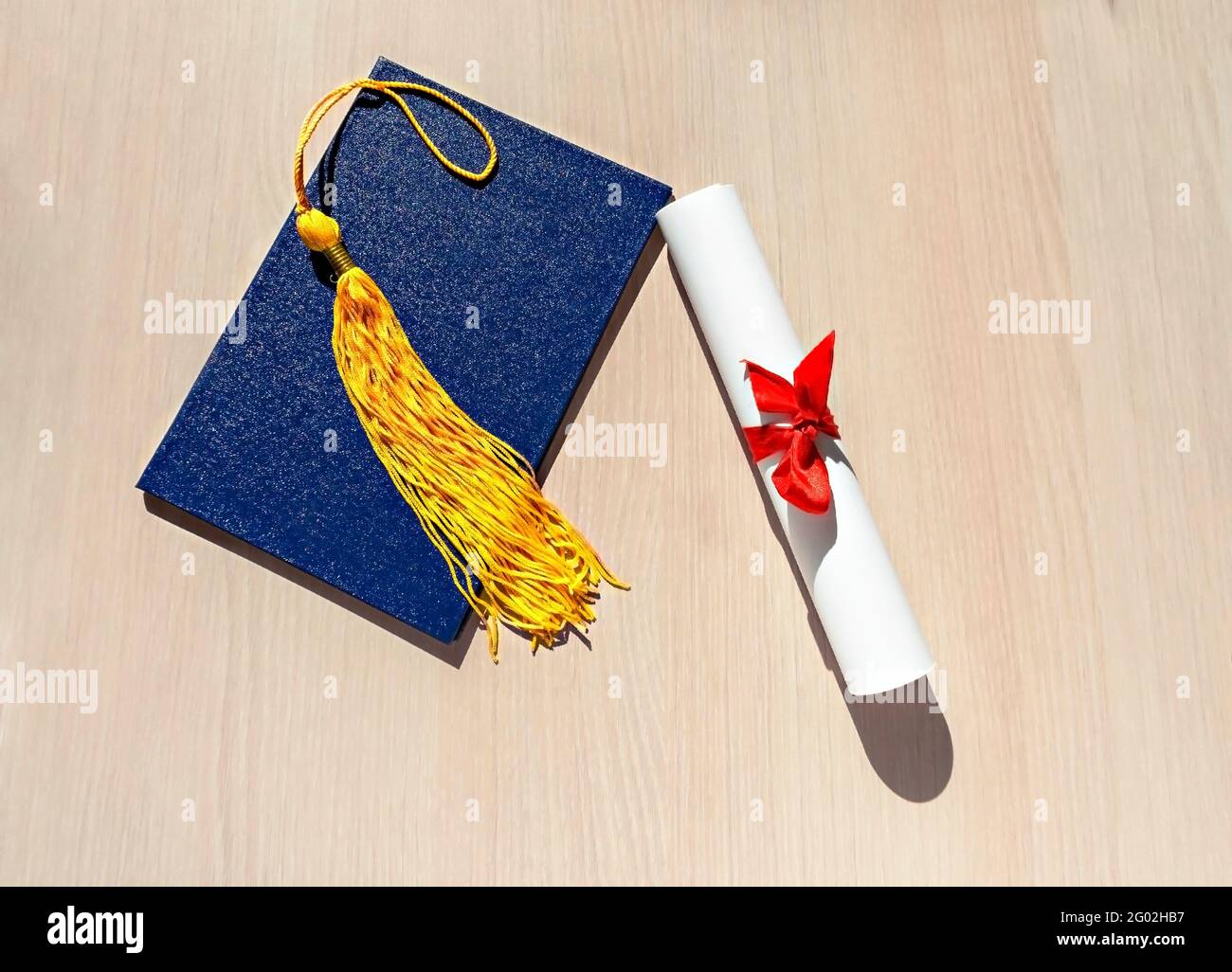 Yellow tassel from graduation cap, blue diploma and paper scroll tied with red ribbon with bow on beige wooden background, Flat lay, top view, mortarb Stock Photo