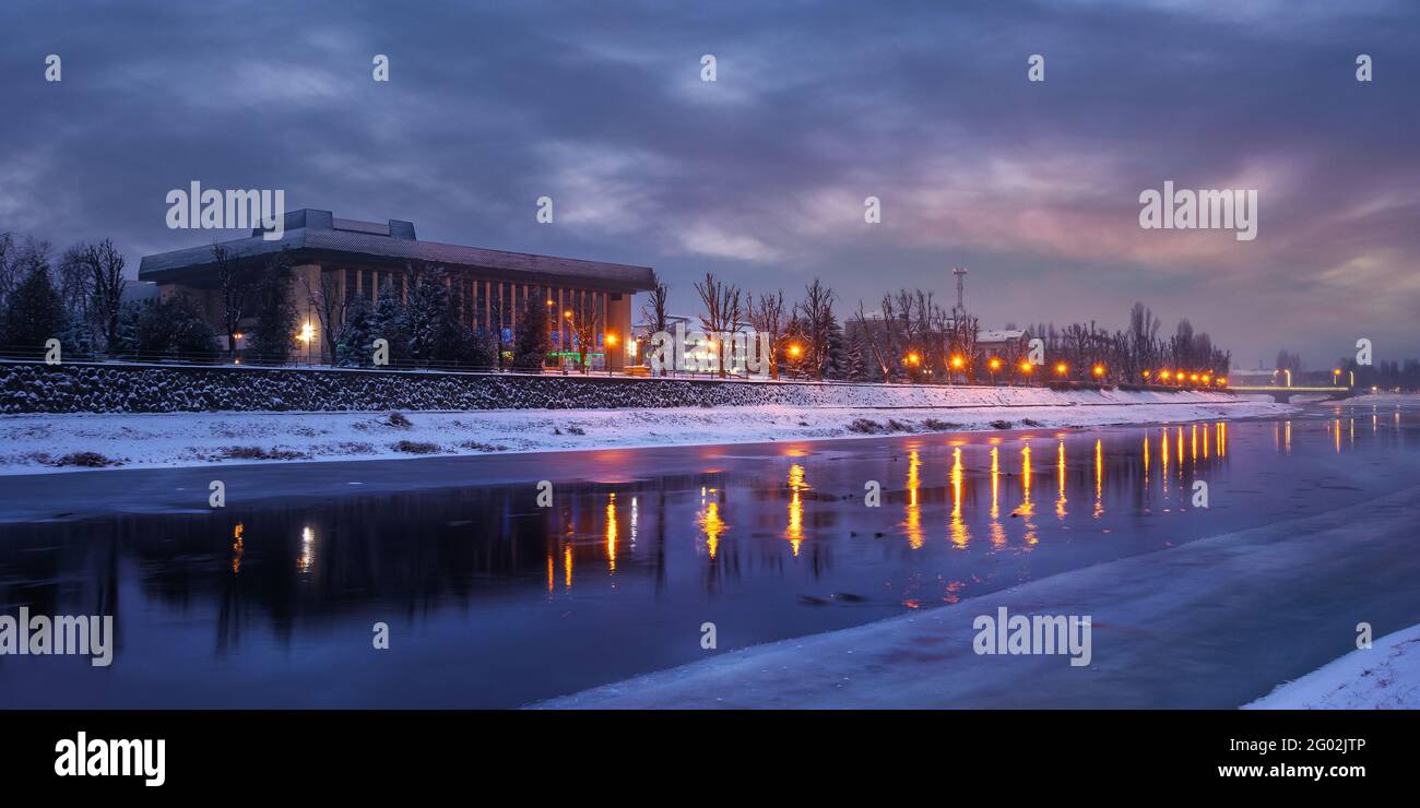 uzhhorod, ukraine - 26 DEC 2016: winter cityscape at dawn. beautiful scenery on the river uzh. city lights reflecting in the water. snow on the embank Stock Photo